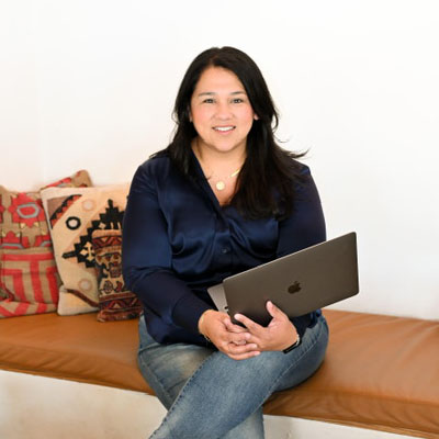 A woman sitting on a leather bench, wearing a navy blouse and blue jeans, holding a laptop and smiling, with colorful patterned pillows in the background.
