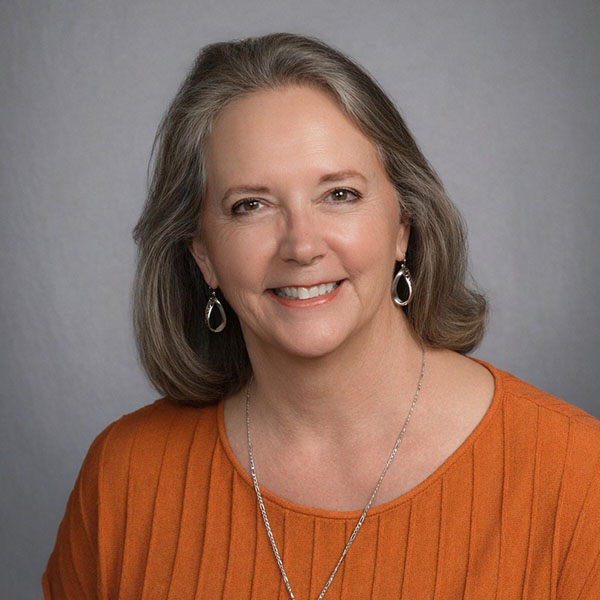 A woman with shoulder-length gray hair smiles warmly at the camera, wearing an orange top and silver dangling earrings. The background is a soft gray.