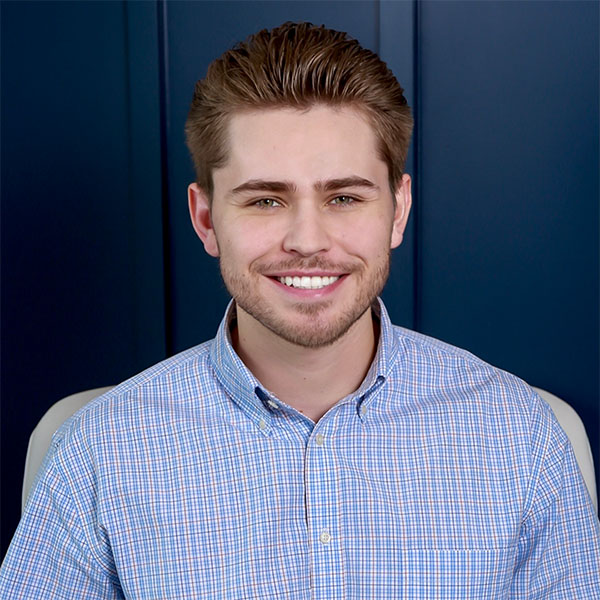 A young man with light brown hair and a well-groomed beard smiles warmly at the camera, wearing a blue checkered button-up shirt, seated in front of a dark blue background.